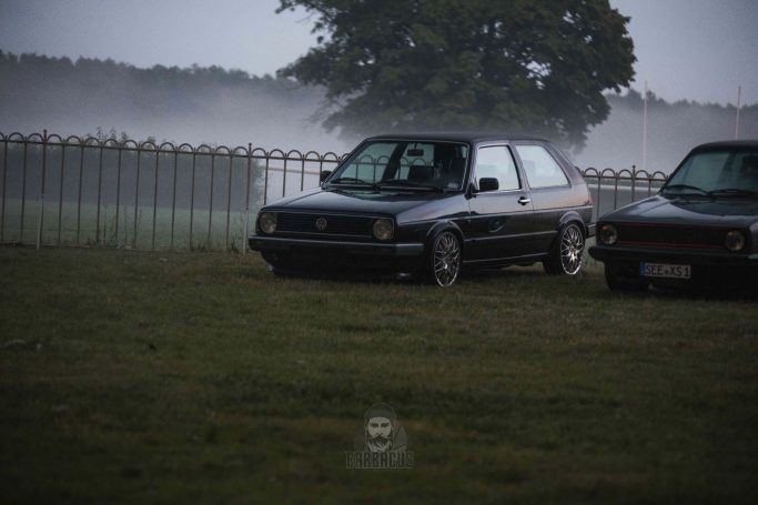 Schwarzes Auto parkt auf einem Feld bei nebligem Wetter, umgeben von einer Hecke.